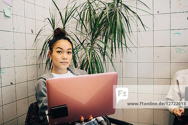 Portrait of confident young female hacker using laptop while sitting against tile wall at small creative office