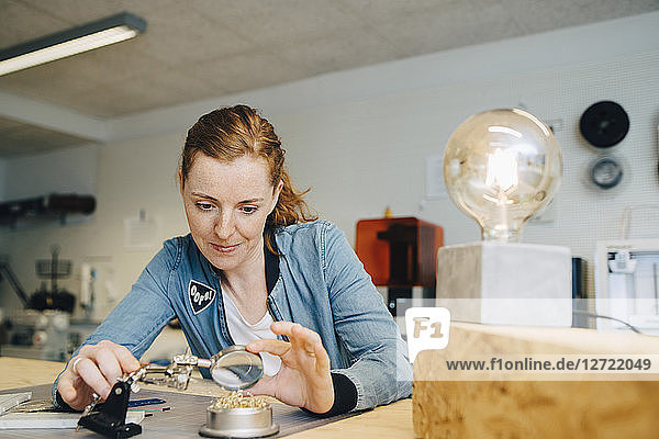 Confident redhead female engineer looking through magnifying glass at workbench in creative office