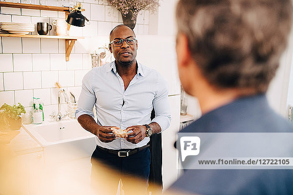 Man holding bread slice while talking to friend in kitchen at home