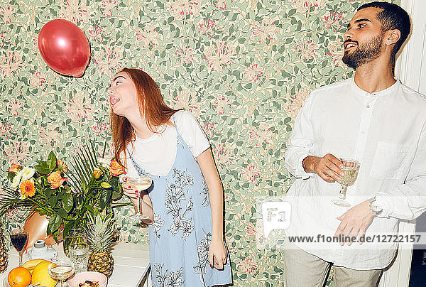 Young man looking at woman playing with balloon while standing against wallpaper at home during dinner party