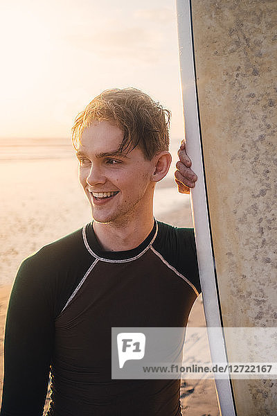 Smiling young man with surfboard standing at beach during sunset