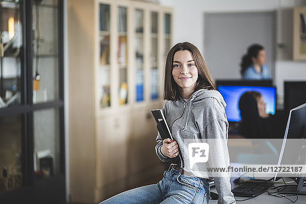 Portrait of smiling high school female student sitting with books on desk in classroom