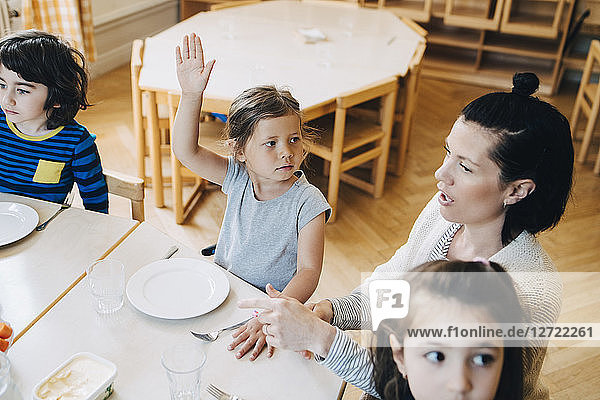 High angle view of girl with arm raised sitting amidst teacher and friends at table in classroom