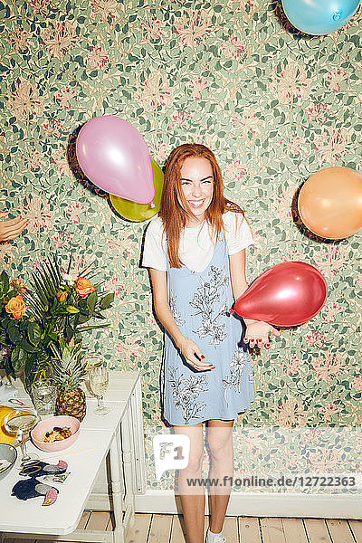 Portrait of cheerful young woman standing amidst balloons by table against wallpaper during party at home