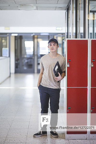 Portrait of confident teenage boy holding book while standing by lockers in corridor at high school