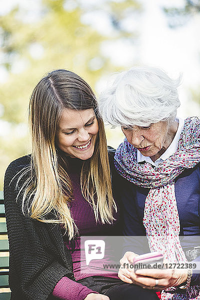 Senior woman showing mobile phone to granddaughter while sitting on bench outdoors