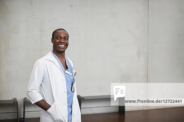 Portrait of smiling male doctor standing with hands in pockets against wall at hospital