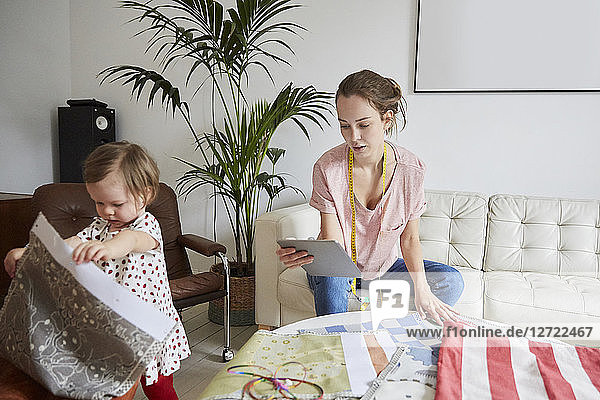 Fashion designer with digital tablet examining textile while daughter playing at home
