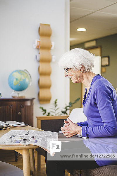 Side view of senor woman smiling while reading newspaper at table in nursing home