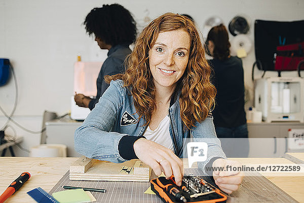 Portrait of smiling redhead female technician sitting at workbench while colleagues working in creative office