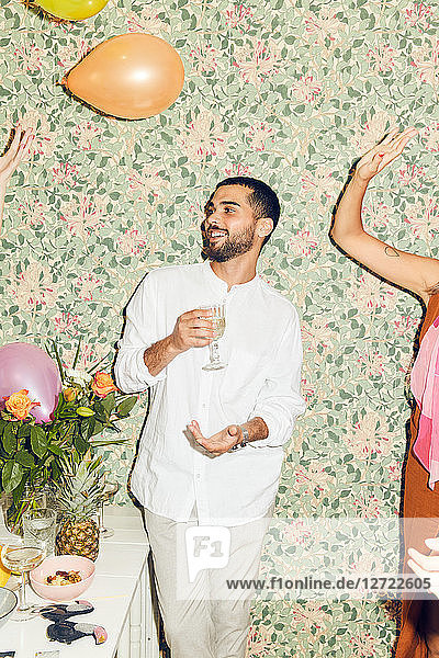 Smiling young man holding drink while standing by woman playing with balloons against wallpaper at home during dinner pa