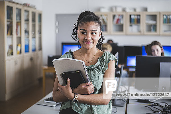 Portrait of smiling teenage girl standing with book in computer lab at high school