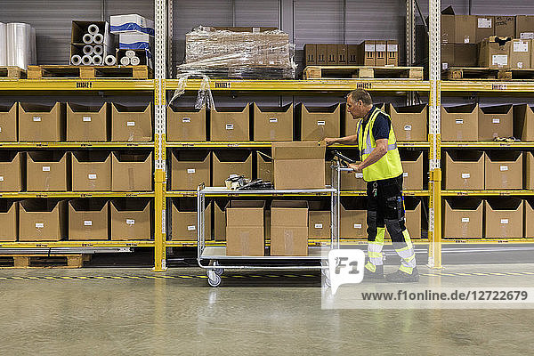 Full length side view of senior male worker pushing cart with cardboard boxes by rack at distribution warehouse