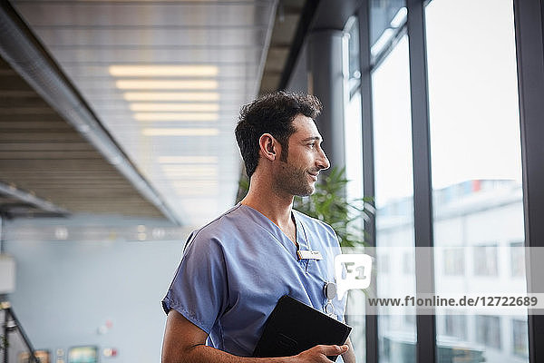 Thoughtful young male nurse holding digital tablet while looking through window at hospital