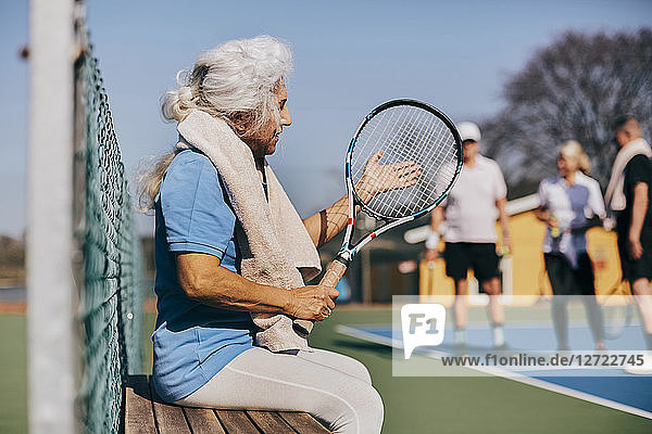 Senior woman holding tennis racket while sitting on bench at tennis court
