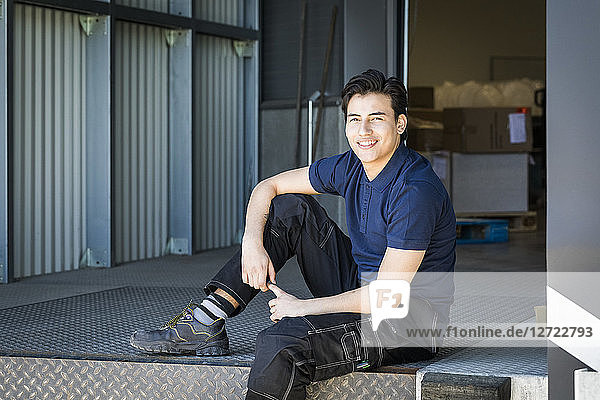 Portrait of smiling young male worker sitting on warehouse entrance