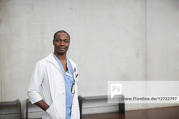 Portrait of confident male doctor standing with hands in pockets against wall at hospital