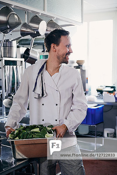 Young male chef smiling while holding container of leaf vegetable in kitchen
