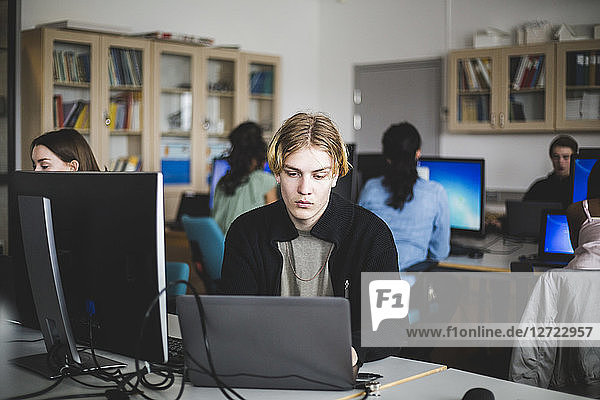 Young male high school student using laptop at desk against teacher and friends sitting in computer lab