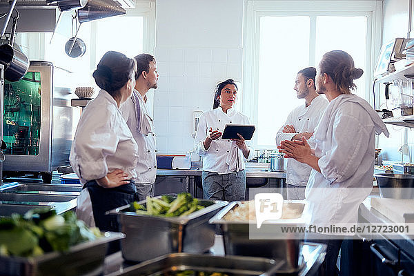 Mid adult female chef holding digital tablet while discussing with team in kitchen