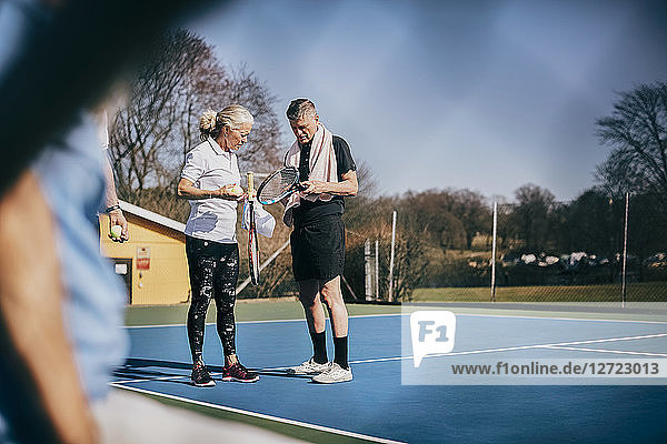 Senior man and woman with rackets talking while standing at tennis court