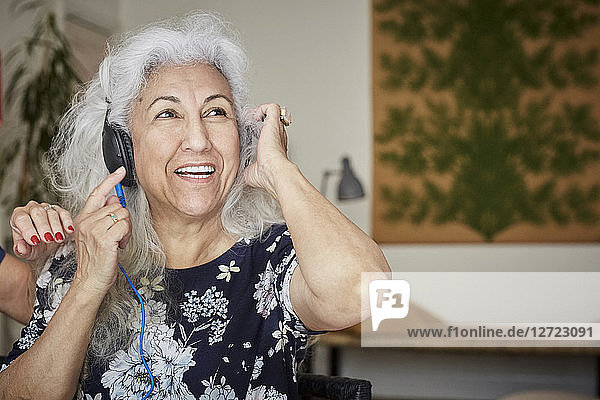 Smiling senior woman wearing headphones looking away while sitting with friend at home