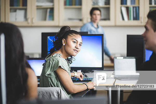 Confident female high school student looking at friend sitting in computer lab