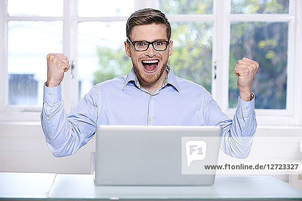Mann in blauem Hemd  Brille  Bart  lächelnd  Fenster im Hintergrund unscharf  Mund geöffnet; Hände in der Luft; sitzend  auf seinen Laptop blickend  frontal .