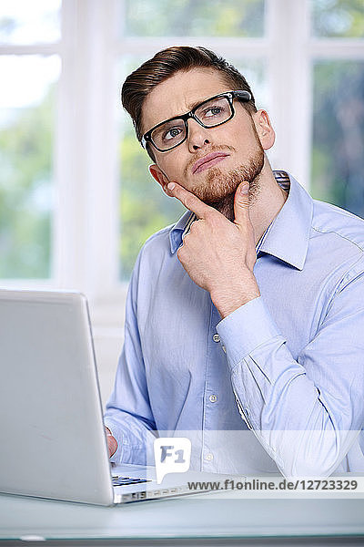 Mann in blauem Hemd  Brille  Bart  ernst  unscharfes Fenster im Hintergrund  sitzend  vor einem Laptop; Blick nach oben  2 Finger am Kinn  Blick nach oben.