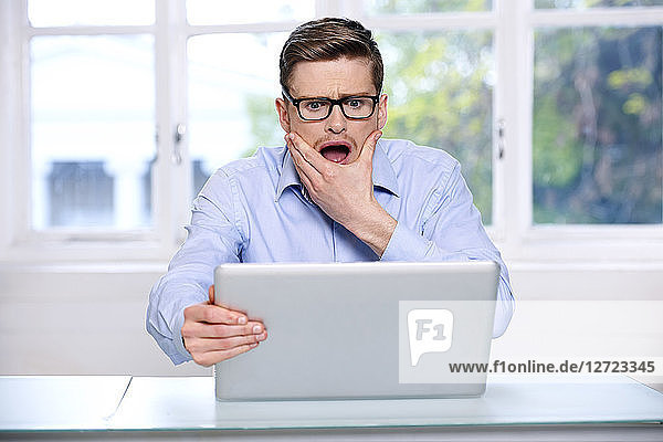 Mann in blauem Hemd; Brille; Bart; ernst; unscharfes Fenster im Hintergrund; geöffneter Mund; Hand im Gesicht; sitzend; Blick auf seinen Laptop; ängstlicher Blick