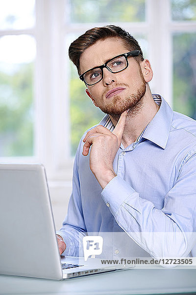 Mann in blauem Hemd  Brille  Bart  ernst  unscharfes Fenster im Hintergrund  sitzend  vor einem Laptop; Blick nach oben  Finger unter dem Kinn  Blick nach oben.