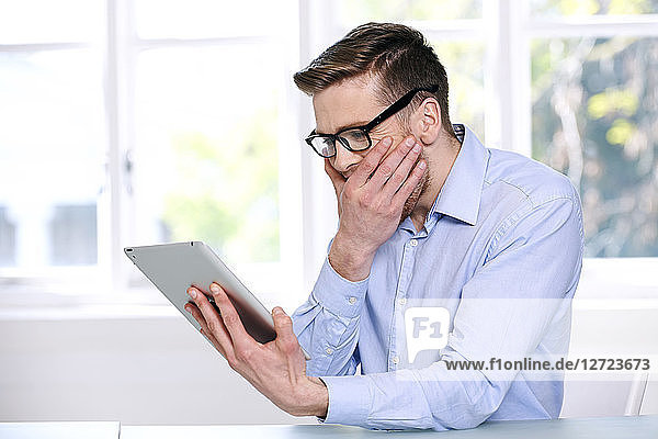 Mann in blauem Hemd; Brille; Bart; lächelnd; Fenster im Hintergrund unscharf; sitzend; Blick auf Tablet  Hand auf dem Mund .