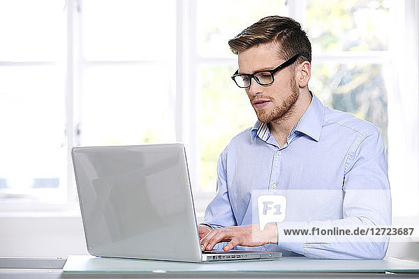 man in a blue shirt  glasses  beard  serious  window out of focus in the background  sitting  typing on a computer laptop