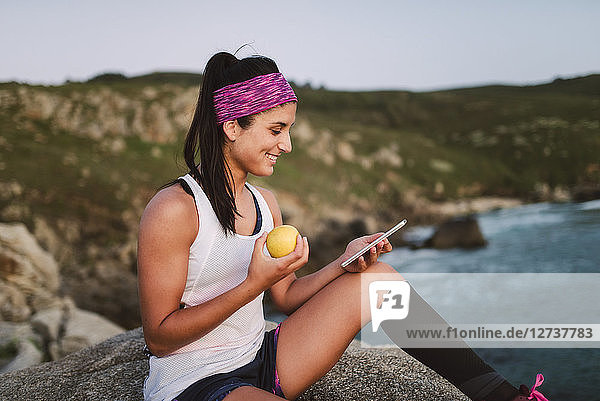 Sportive woman sitting on rocks in the evening  holding apple and using smartphone
