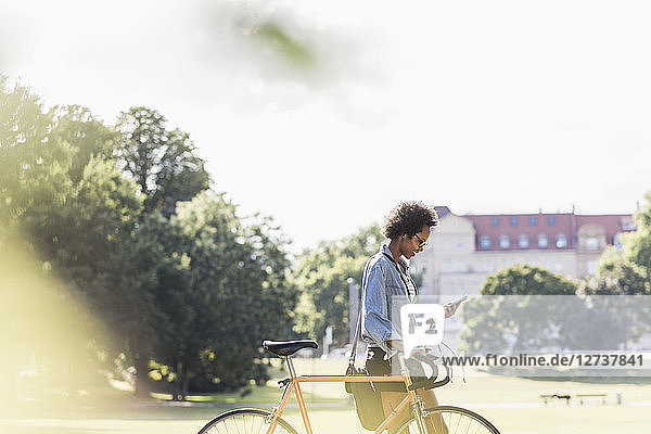 Young woman with cell phone pushing bicycle in park