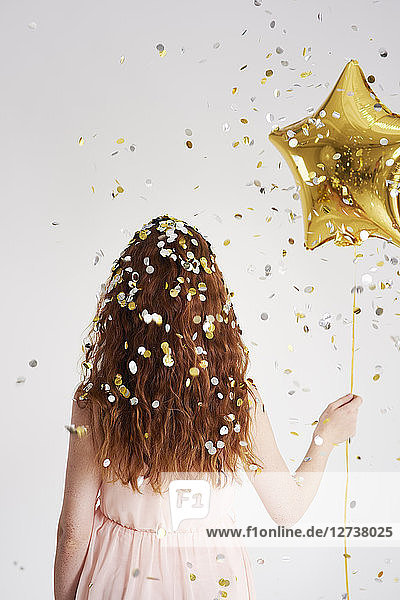 Back view of redheaded young woman with star-shaped golden balloon under shower of confetti