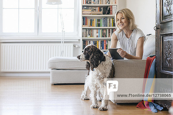 Smiling mature woman with dog lying on couch at home holding tablet