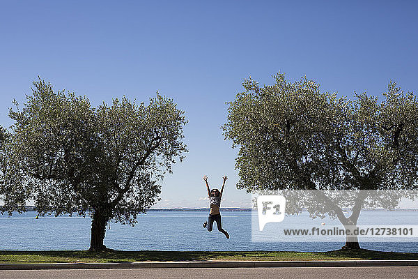 Italy  Lake Garda  happy young woman jumping in the air between two trees
