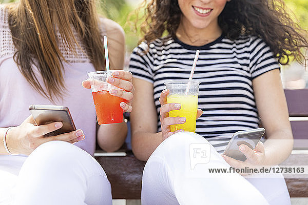 Close-up of two female friends sitting on a bench with slush and cell phones
