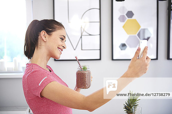 Smiling young woman with smoothie taking selfie with smartphone in the kitchen