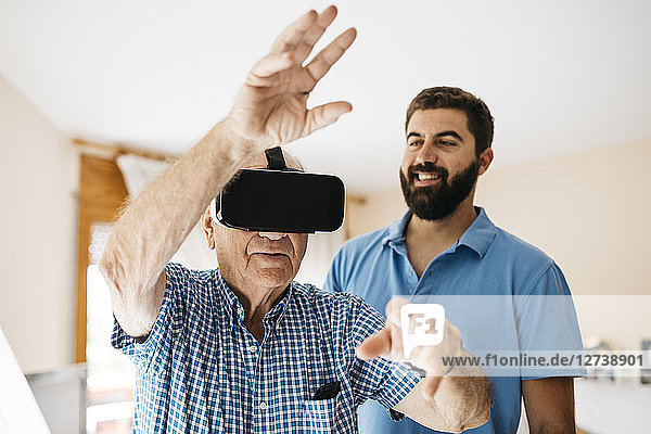 Senior man using Virtual Reality Glasses at home while his adult grandson watching him