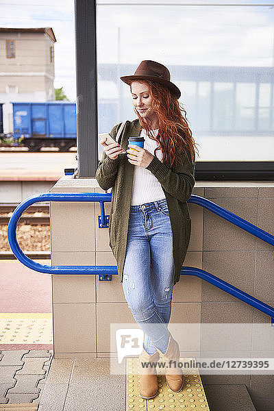 Smiling redheaded woman with coffee to go looking at cell phone at platform