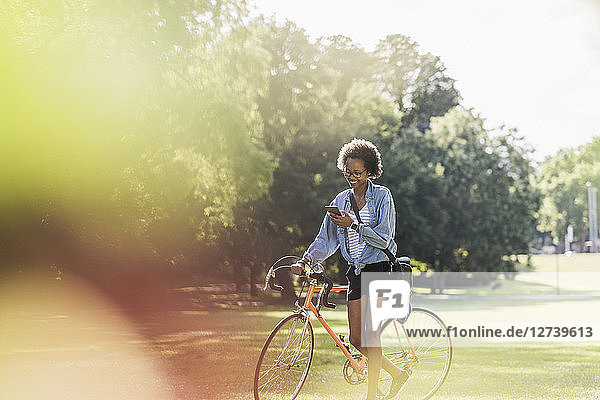 Young woman with cell phone pushing bicycle in park
