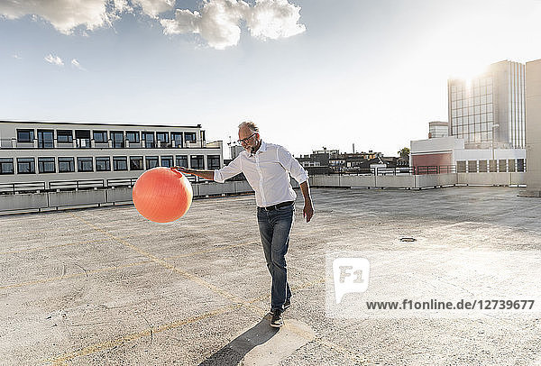 Mature man playing with orange fitness ball on rooftop of a high-rise building