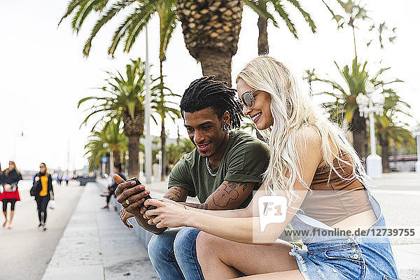 Spain  Barcelona  multicultural young couple sitting beside promenade looking at cell phone