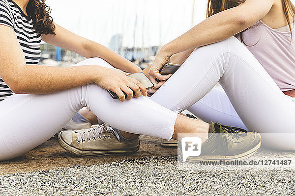 Close-up of two female friends sitting on ground holding cell phones