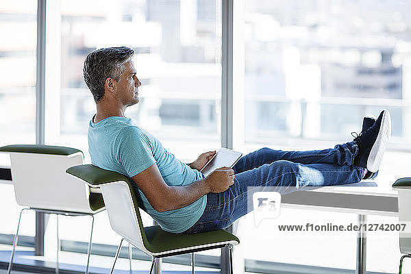 Businessman sitting in office with feet up  holding digital tablet
