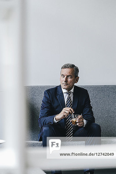 Mature businessman with glass of coffee sitting on couch in his office looking out of window
