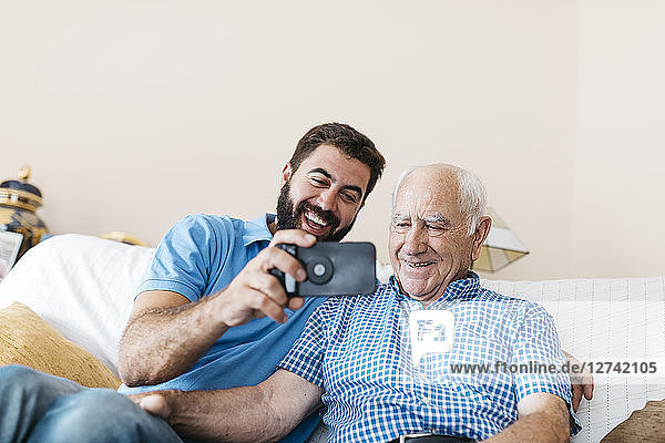 Portrait of adult grandson and his grandfather taking selfie with smartphone at home