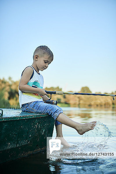 Little boy with fishing rod sitting on boat splashing with water